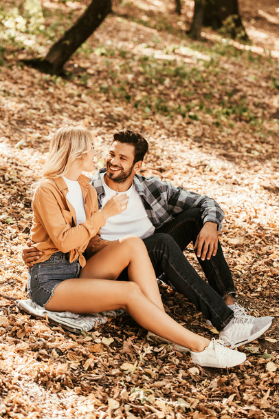 happy young couple looking at each other while sitting on fall foliage in park - Foto, Imagem