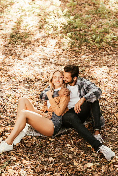 happy man embracing cheerful girlfriend while sitting on fall foliage in park - Photo, image