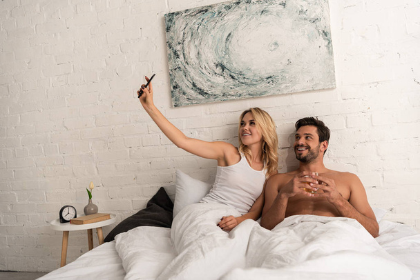 hermosa pareja sonriente tomando selfie en el teléfono inteligente en la cama por la mañana
 - Foto, Imagen