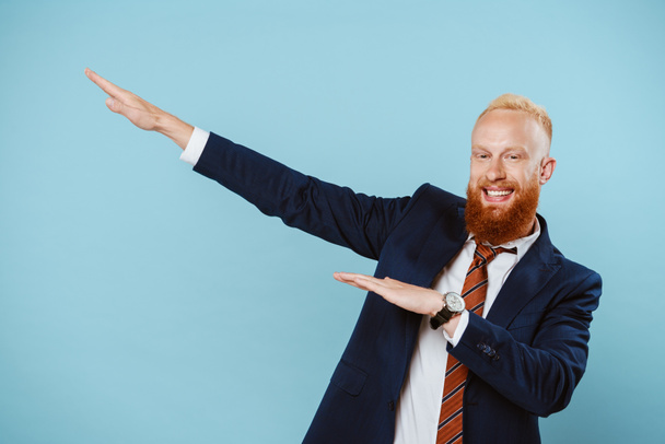 cheerful bearded businessman in suit gesturing isolated on blue - 写真・画像