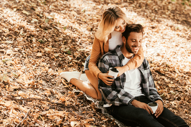 happy girl embracing cheerful boyfriend while sitting on fall foliage in park - Photo, image