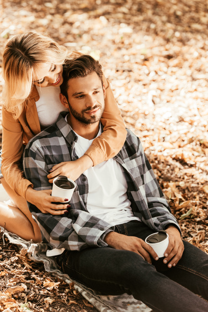 happy girl embracing dreamy boyfriend while sitting on fall foliage in park - Valokuva, kuva