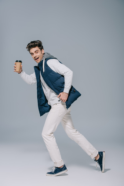 stylish smiling man in warm waistcoat running with coffee to go on grey - Фото, зображення