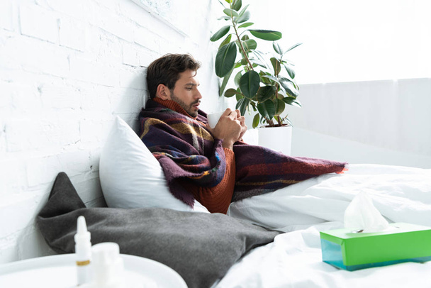 ill man with cup of hot drink lying in bed with napkins and nasal spray - Foto, immagini