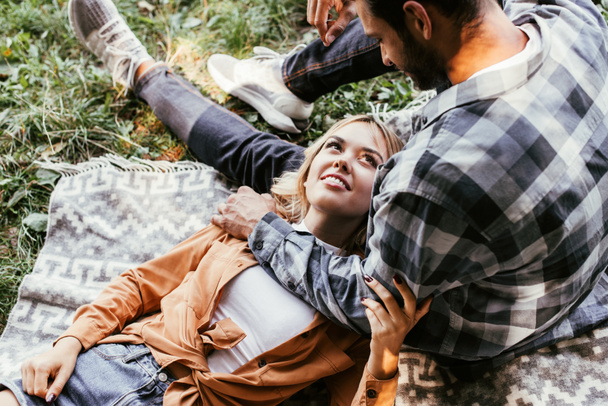 happy young woman lying on blanket in park near sitting boyfriend - Photo, Image