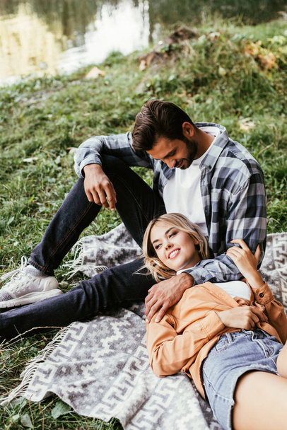 handsome man embracing happy girlfriend lying on blanket near lake in park - Photo, Image