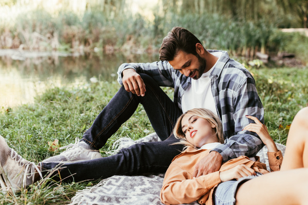 handsome man embracing girlfriend sleeping on blanket near lake in park - Fotografie, Obrázek