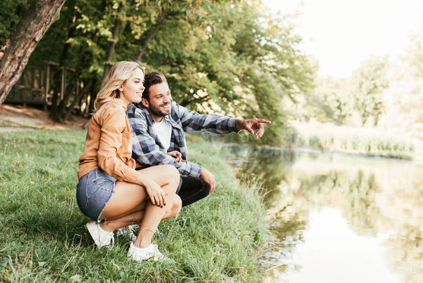 cheerful man pointing with finger while squatting near lake with girlfriend - Фото, изображение