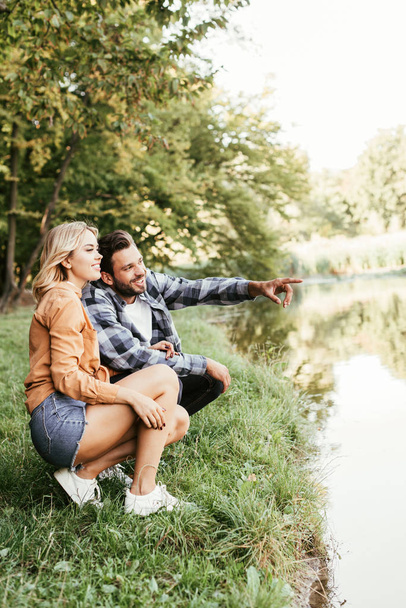 smiling man pointing with finger while squatting near lake with girlfriend - Fotoğraf, Görsel