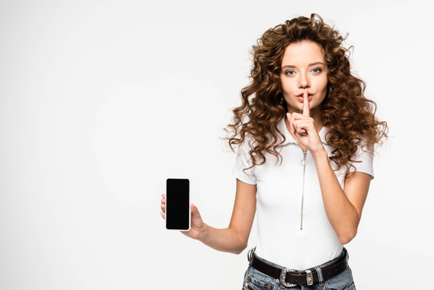 σγουρό κορίτσι δείχνει το σύμβολο της σιωπής και smartphone με λευκή οθόνη, που απομονώνονται σε λευκό - Φωτογραφία, εικόνα