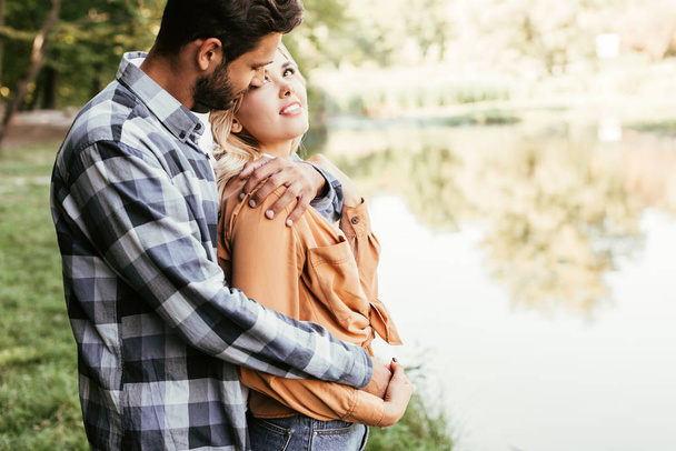 handsome man embracing happy girlfriend while standing near lake in park - Photo, Image