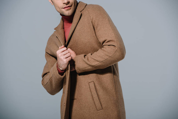 καλλιεργημένη άποψη της μόδας άνθρωπος ποζάρουν σε μπεζ παλτό, απομονώνονται σε γκρι - Φωτογραφία, εικόνα