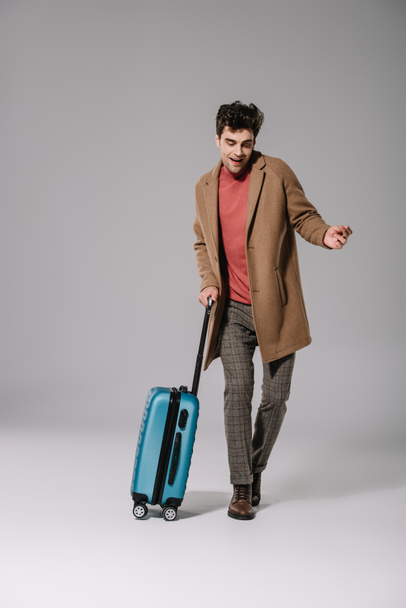 χαρούμενος κομψός άντρας σε μπεζ παλτό χορεύει με ταξιδιωτική τσάντα σε γκρι - Φωτογραφία, εικόνα