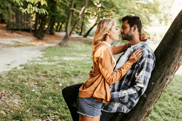 attractive young woman kissing boyfriend while standing tree trunk in park - Фото, зображення