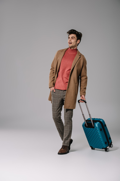 excited man in beige coat walking with suitcase on grey - Фото, изображение