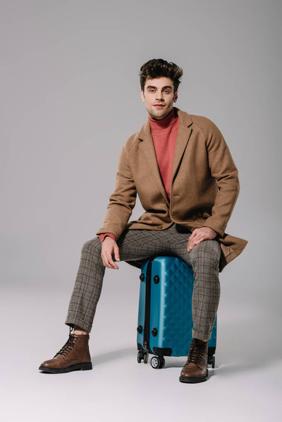 handsome man in beige coat sitting on suitcase on grey - Foto, afbeelding