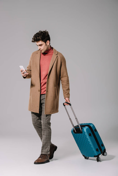 stylish man in beige coat with travel bag using smartphone on grey - Foto, afbeelding
