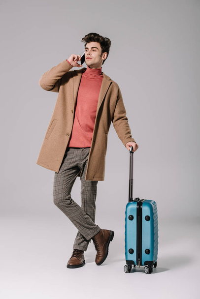 fashionable man in beige coat talking on smartphone and holding travel bag on grey - Фото, изображение
