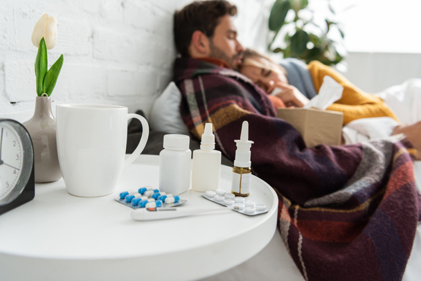 selectieve focus van zieke liggend in bed met thermometer, warme drank en neusspray en pillen op tafel  - Foto, afbeelding