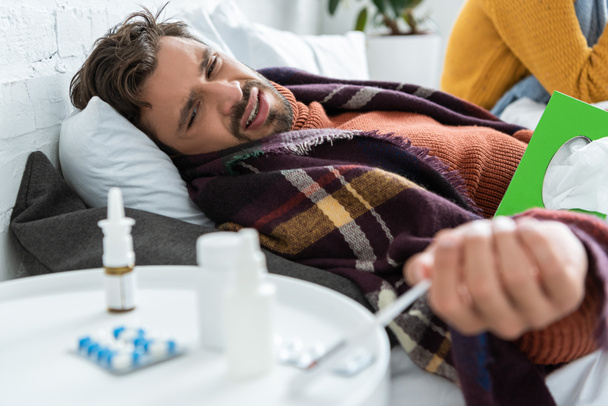 homem doente na cama com pílulas, guardanapos e spray nasal no quarto com mulher
 - Foto, Imagem