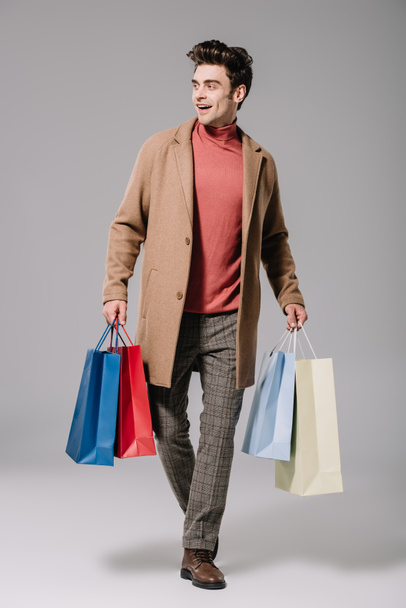 happy fashionable man in beige coat walking with shopping bags on grey - Photo, Image