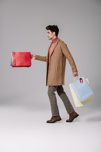 side view of stylish man in beige coat walking with shopping bags on grey - Foto, immagini