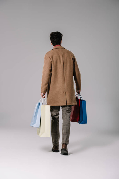 rear view of man in beige coat holding shopping bags on grey - Фото, изображение