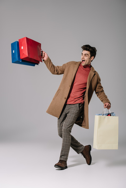 excited stylish man in beige coat walking with shopping bags on grey - Фото, зображення