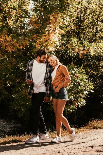 happy young woman leaning on shoulder of boyfriend while walking in park - Photo, Image