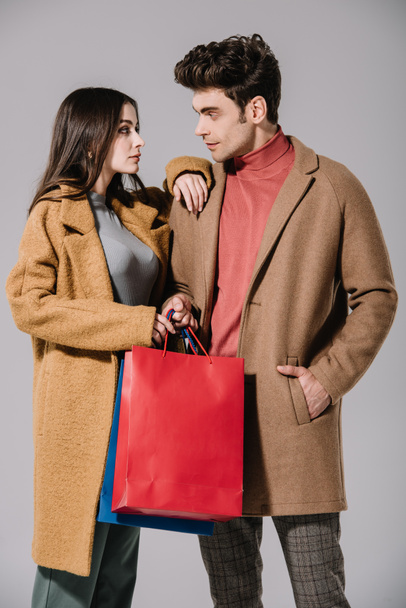 ζευγάρι σε μπεζ παλτά με τσάντες αγορών να κοιτάζονται, απομονωμένο σε γκρι - Φωτογραφία, εικόνα