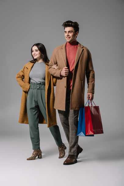 smiling couple in beige coats posing with shopping bags on grey - Фото, зображення