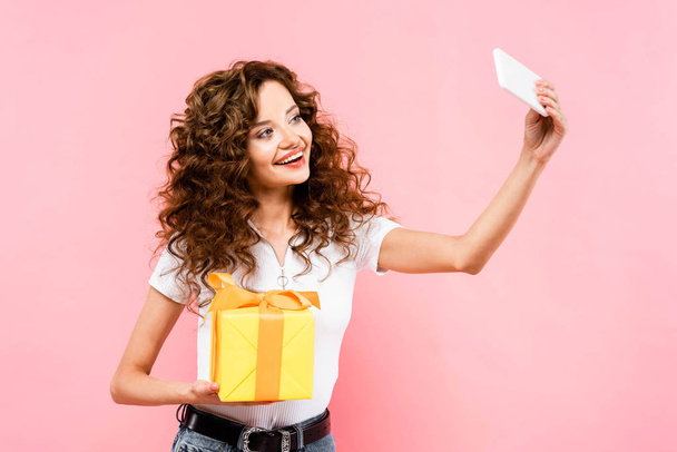attractive happy curly girl taking selfie with gift box, isolated on pink - 写真・画像