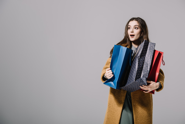 attractive surprised girl in beige coat holding shopping bags, isolated on grey - Fotografie, Obrázek