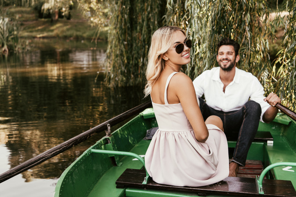 cheerful young woman looking at camera while sitting in boat near happy boyfriend - Fotografie, Obrázek