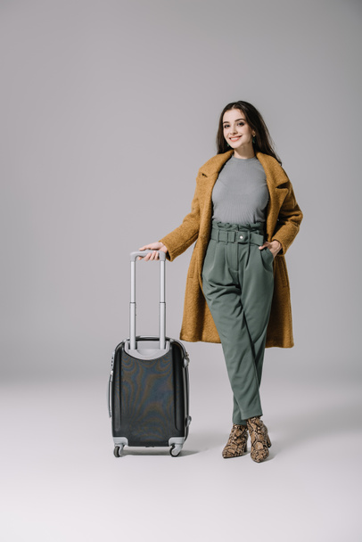 beautiful smiling girl in beige coat posing with travel bag on grey - Фото, изображение