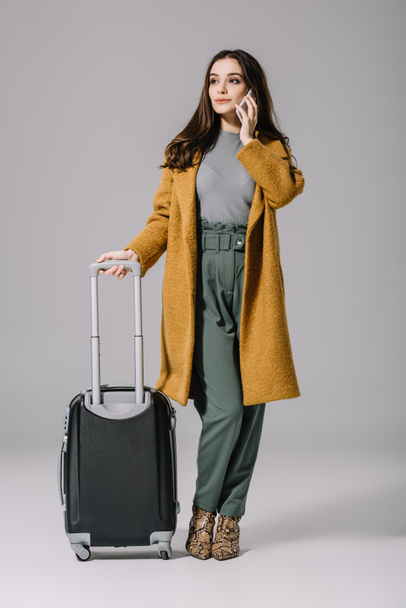 beautiful stylish girl in beige coat standing with travel bag while talking on smartphone on grey - Fotó, kép
