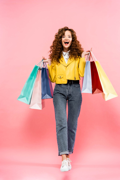 excited curly girl holding shopping bags, isolated on pink - Foto, afbeelding