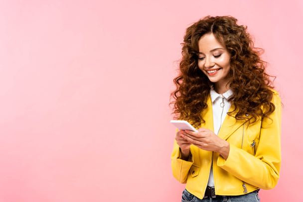 cheerful curly girl using smartphone, isolated on pink - Foto, imagen