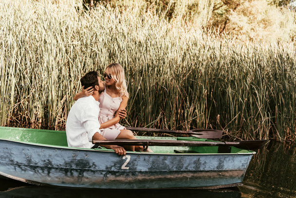 young couple hugging and kissing in boat on river near thicket of sedge - Фото, изображение