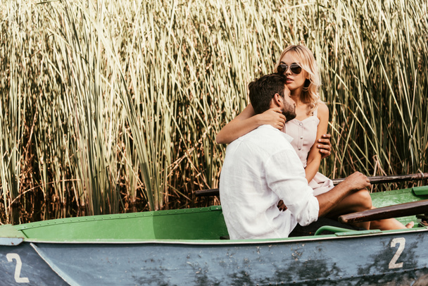 attractive young woman embracing boyfriend in boat on lake near thicket of sedge - Foto, imagen