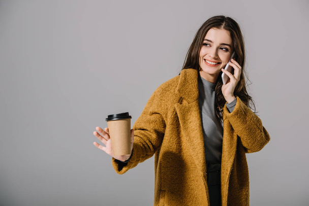 smiling girl in beige coat holding coffee to go while talking on smartphone, isolated on grey - Foto, Imagem