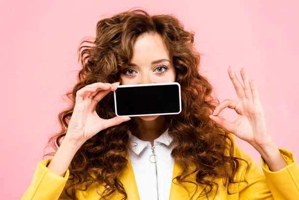 curly girl showing ok sign and smartphone with blank screen, isolated on pink - Photo, Image
