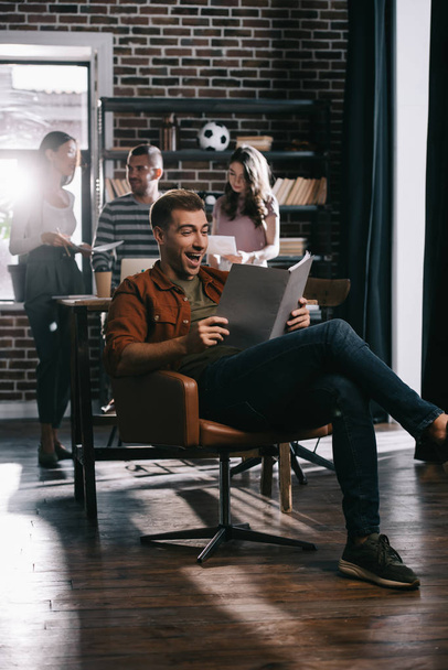 cheerful businessman sitting in armchair and reading document near young colleagues - Foto, Imagem