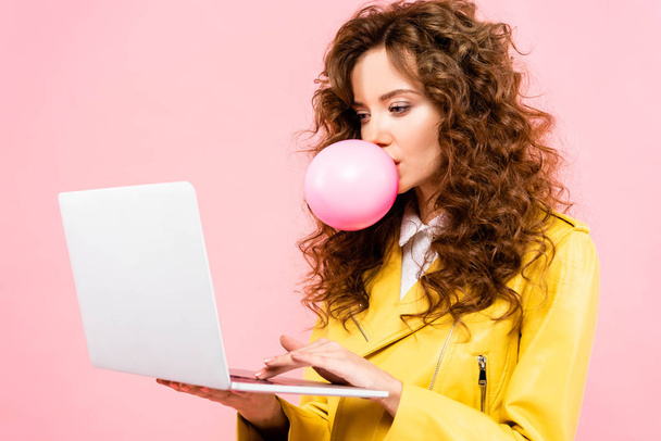 šťastná kudrnatá žena s bublinou žvýkačky pomocí notebooku, izolované na růžové - Fotografie, Obrázek