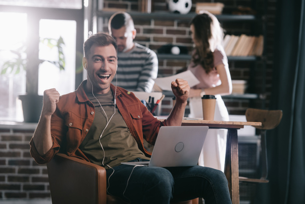 cheerful businessman in earphones sitting in armchair, using laptop and showing winner gesture near young colleagues - Fotó, kép
