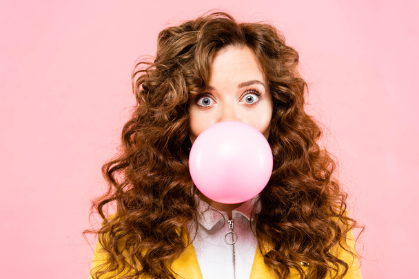 atraktivní překvapená kudrnatá dívka s bublinkovou žvýkačkou, izolovaná na růžové - Fotografie, Obrázek