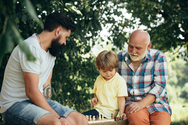 Cute little boy playing chess with Parents. Three different generations ages: grandfather father and son together. - Photo, image