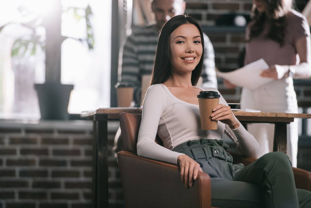 young businesswoman sitting in armchair with coffee to go and smiling at camera near colleagues - Foto, imagen