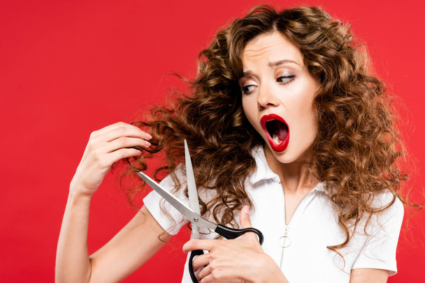 shocked curly girl cutting her hair with scissors, isolated on red - Fotografie, Obrázek