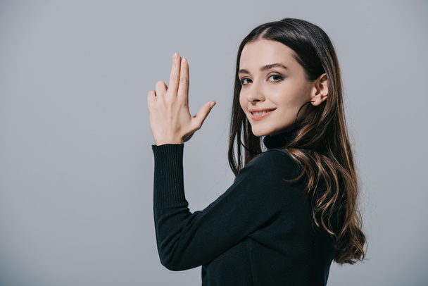 smiling fashionable girl in black sweater showing hand gun, isolated on grey - Foto, Imagem